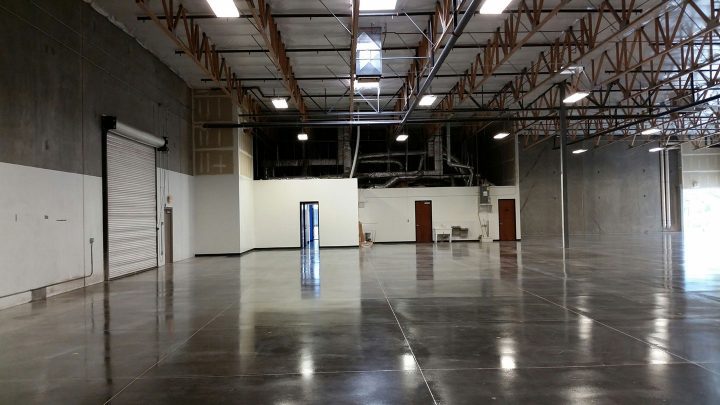 Interior of a spacious, empty industrial home warehouse with glossy concrete floors, exposed ceiling beams, and a rolled-up door on the left.